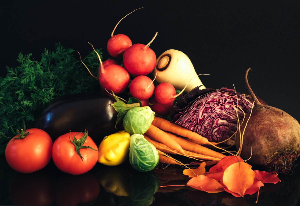 Fresh vegetables piled against a dark background — tomatoes, radishes, carrots, red cabbage, and herbs